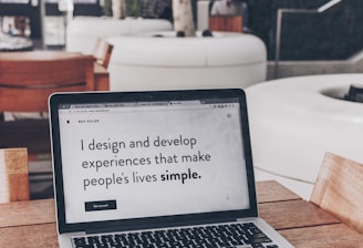 A laptop displaying a webpage with the text 'I design and develop experiences that make people's lives simple.' is placed on a wooden table. It is set in an outdoor seating area with white furniture and greenery in the background. The setting appears to be calm and conducive to work or relaxation.