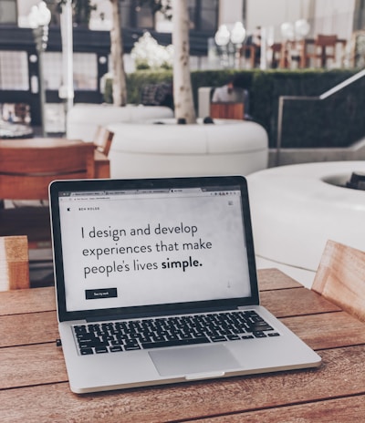 A laptop displaying a webpage with the text 'I design and develop experiences that make people's lives simple.' is placed on a wooden table. It is set in an outdoor seating area with white furniture and greenery in the background. The setting appears to be calm and conducive to work or relaxation.