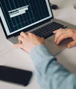 A person is typing on a silver laptop with a screen displaying text about designing digital solutions for big customers. Nearby, there is a black smartphone and white earphones placed on the desk. The person's sleeves are light blue, suggesting a casual attire.
