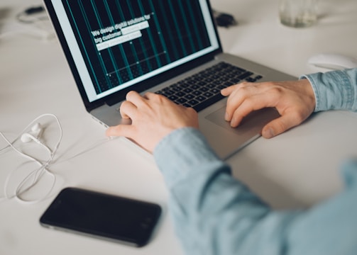 A person is typing on a silver laptop with a screen displaying text about designing digital solutions for big customers. Nearby, there is a black smartphone and white earphones placed on the desk. The person's sleeves are light blue, suggesting a casual attire.