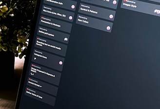 A close-up view of a laptop screen displaying a project management tool with columns labeled 'Open', 'In Progress', and 'Review'. Tasks are listed under each column. The environment appears dimly lit, with a small, leafy plant visible in the background.