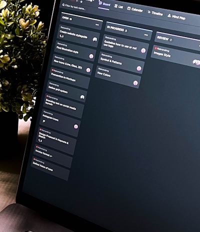A close-up view of a laptop screen displaying a project management tool with columns labeled 'Open', 'In Progress', and 'Review'. Tasks are listed under each column. The environment appears dimly lit, with a small, leafy plant visible in the background.