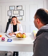 A professional consultation setting with a medical professional sitting at a desk facing a client. The room has a modern aesthetic with white walls decorated with framed certificates. The desk is organized with office supplies, a laptop, and a fruit bowl in the center.