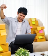 A person is sitting on a couch, animatedly raising a fist in the air with a smile, holding a laptop on their lap. Surrounding them are numerous cardboard boxes with colorful sticky notes attached, suggesting a home-based business setup.