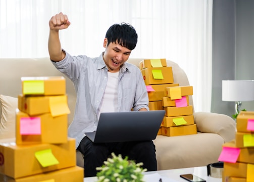 A person is sitting on a couch, animatedly raising a fist in the air with a smile, holding a laptop on their lap. Surrounding them are numerous cardboard boxes with colorful sticky notes attached, suggesting a home-based business setup.